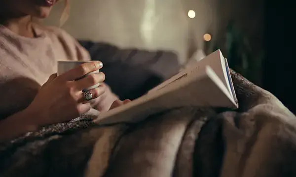 Cropped shot of female hands with book and coffee. Female reading book on bed at night.