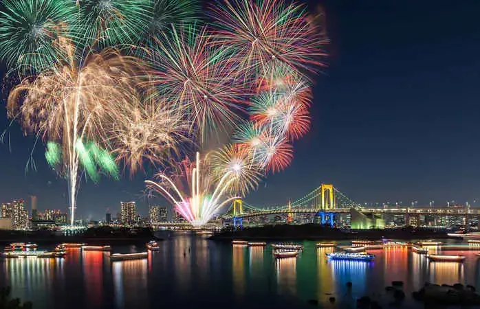 Tokyo's Rainbow bridge is illuminated at dusk with the skyscrapers of Tokyo and fireworks in the distance, Tokyo bay, Japan.
